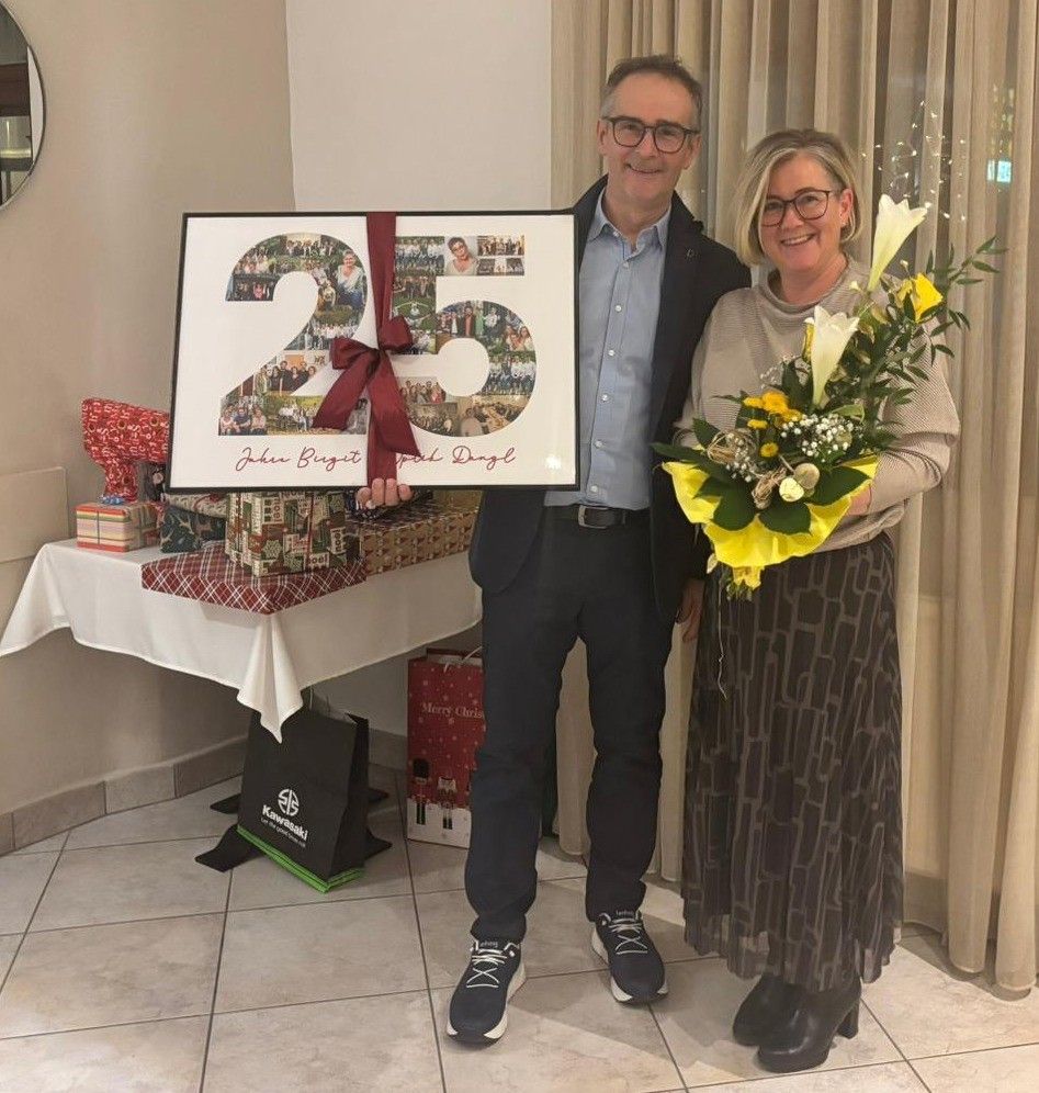 Ein Mann und eine Frau stehen lächelnd in einem Raum. Sie halten eine große, gerahmte Fotocollage mit einer roten Schleife, die die Nummer 25 zeigt. Der Mann trägt eine Brille und einen Gürtel. Hinter ihnen steht ein Tisch mit verpackten Geschenken, der mit einem weißen Tuch bedeckt ist.