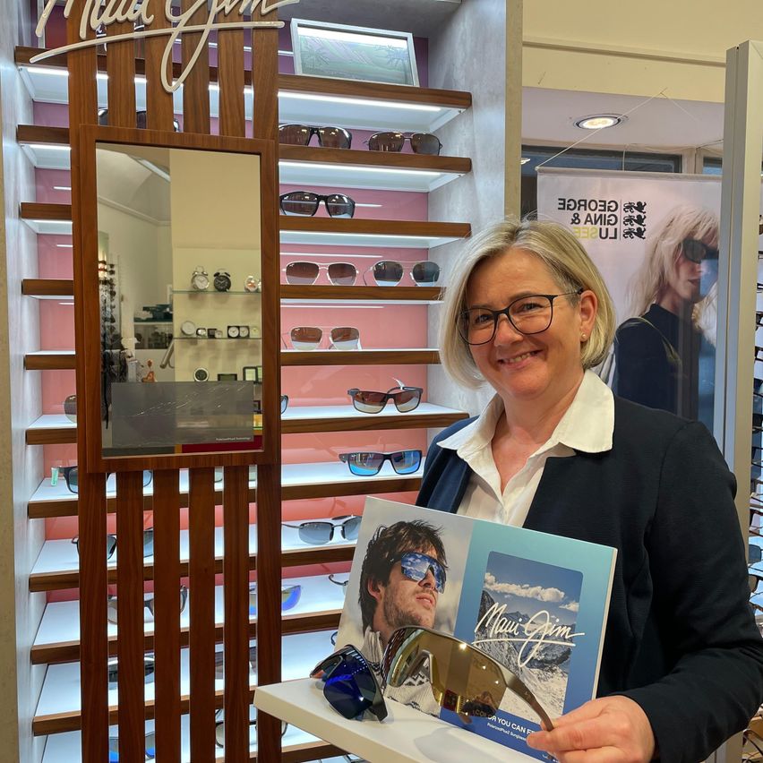 Eine Frau mit Brille steht vor einer Vitrine mit Sonnenbrillen und hält ein Buch und eine Sonnenbrille in den Händen. Dahinter befindet sich ein Regal mit Sonnenbrillen und einem Spiegel.