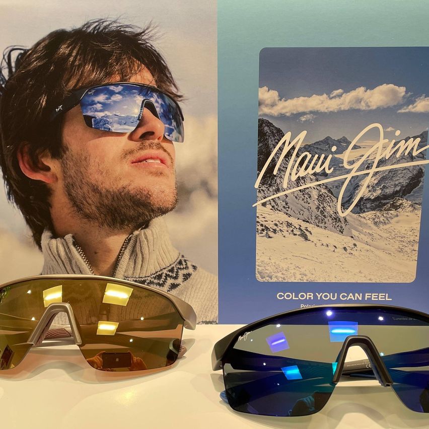 Ein Mann mit reflektierenden Sonnenbrillengläsern, neben einem Poster mit dem Text 'Maui Jim' und 'FARBE, DIE DU SPÜREN KANNST'.