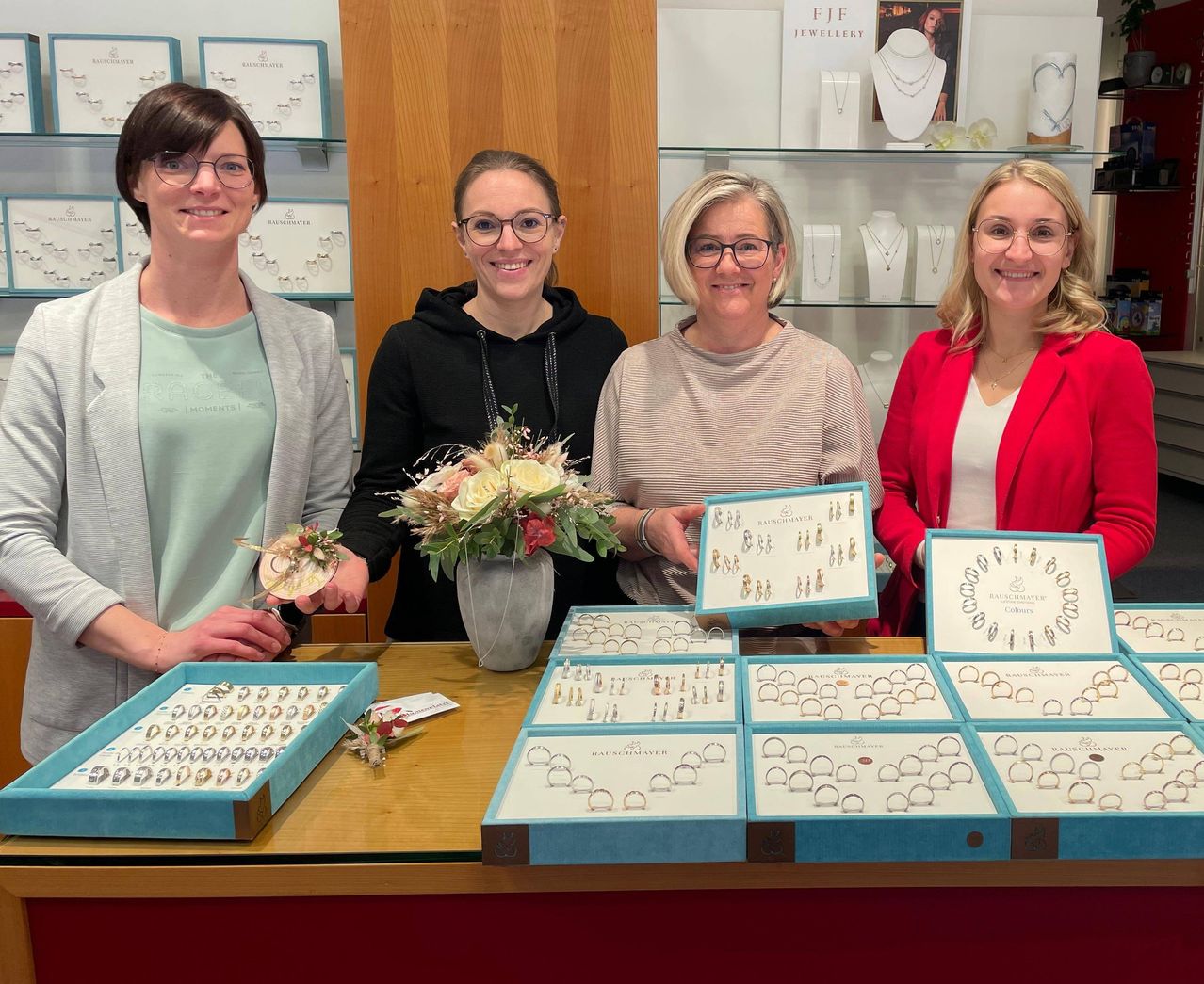 Vier Frauen posieren in einem Juweliergeschäft und lächeln. Dahinter befinden sich Regale mit Schmuckstücken. Auf dem Tisch davor sind verschiedene Schmuckschatullen ausgestellt.