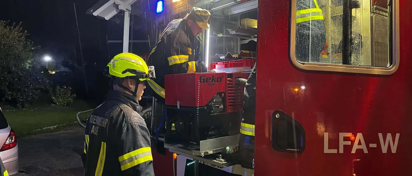 Zwei Feuerwehrleute arbeiten nachts bei Regen an einem roten Feuerwehrauto. Das Auto hat die Buchstaben LFA an der Seite.