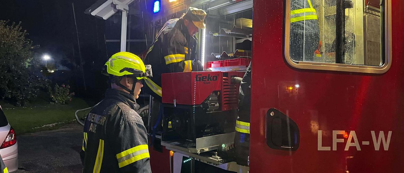 Zwei Feuerwehrleute arbeiten nachts bei Regen an einem roten Feuerwehrauto. Das Auto hat die Buchstaben LFA an der Seite.