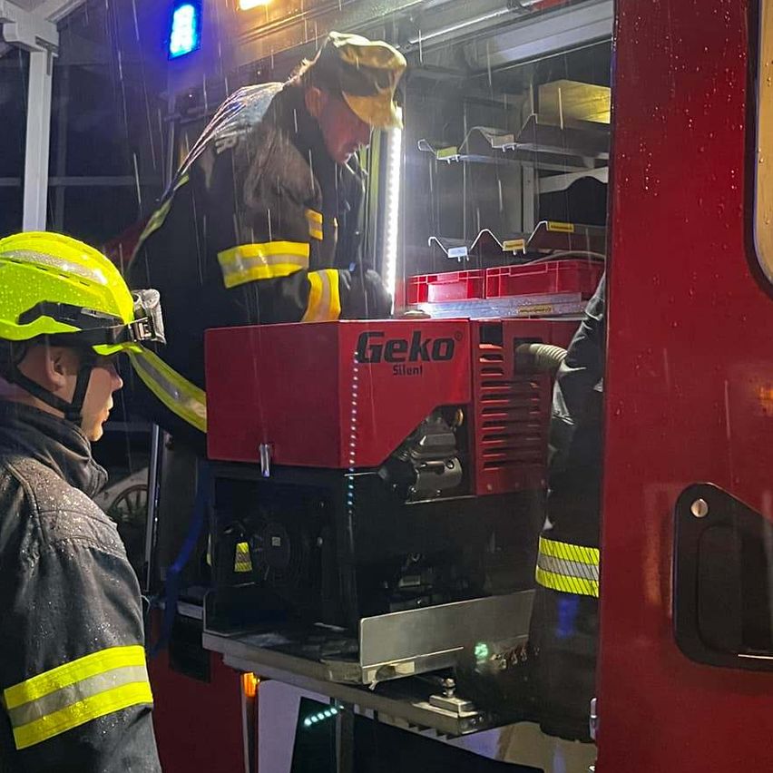 Zwei Feuerwehrleute arbeiten nachts bei Regen an einem roten Feuerwehrauto. Das Auto hat die Buchstaben LFA an der Seite.