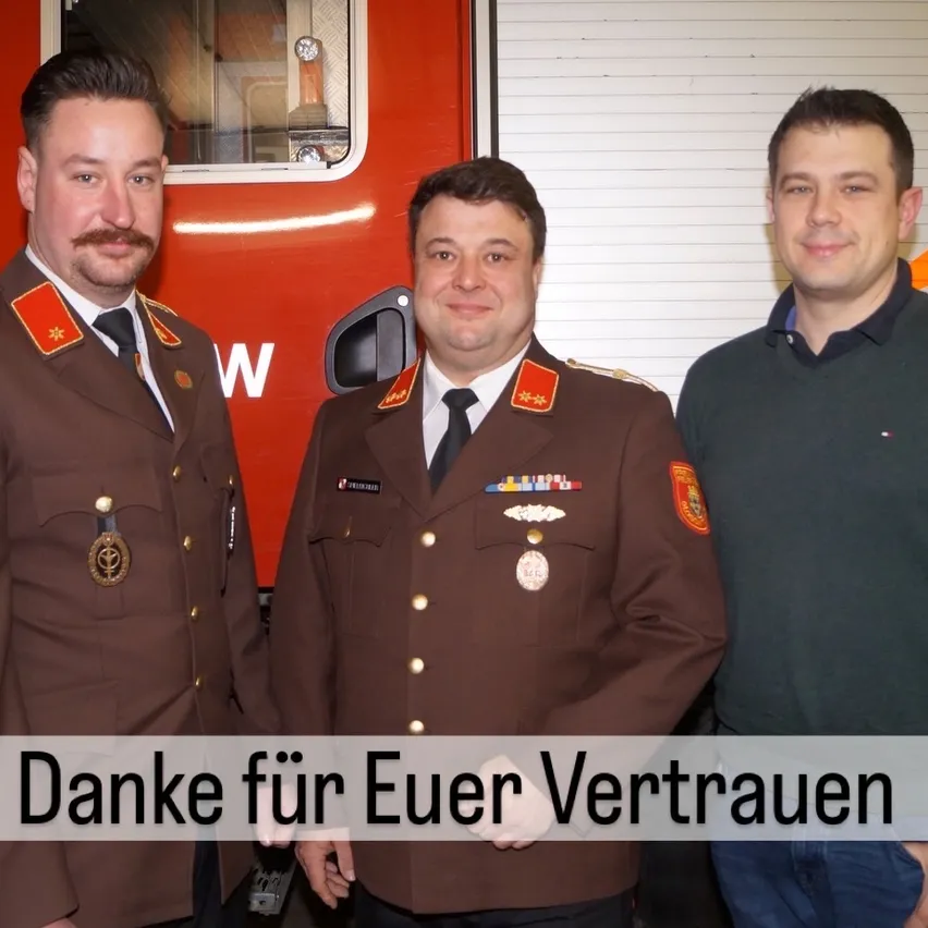 Drei Männer stehen vor einem roten Feuerwehrauto. Der Mann links trägt eine Militäruniform mit Medaillen, der Mann in der Mitte trägt eine braune Uniform mit Medaillen und der Mann rechts trägt einen grünen Pullover. Der Text lautet 'Danke für Euer Vertrauen.'