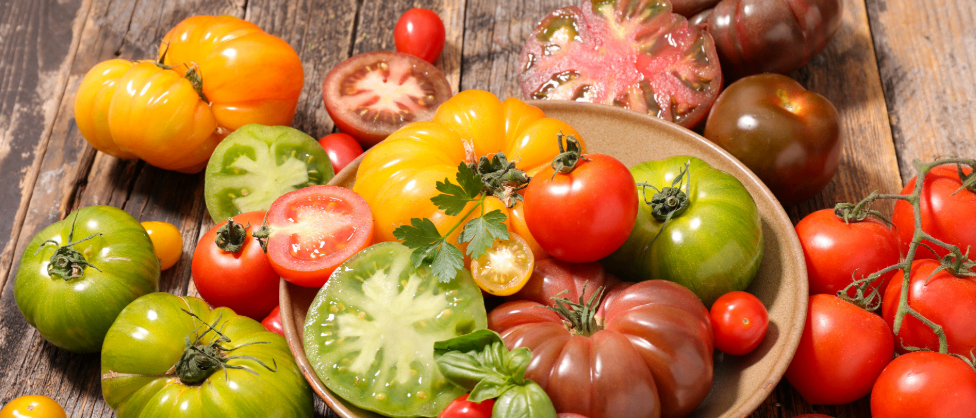 Auf einem Holztisch sind verschiedene Tomaten und Basilikumblätter zu sehen. Im Hintergrund gibt es eine Ankündigung für den Bauernmarkt in Krottendorf am 11. Juli.