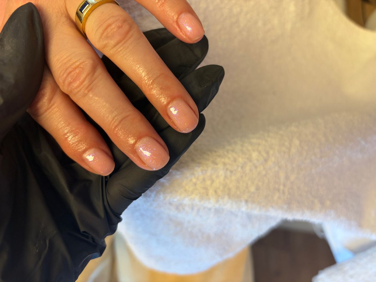Eine Hand mit schwarzen Handschuhen hält gepflegte Nägel mit hellem pinkem Lack. Ein goldener Ring mit einem blauen Stein schmückt den Ringfinger.