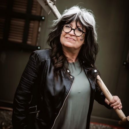 Eine Frau mit weißen Haaren trägt eine schwarze Lederjacke und hält einen hölzernen Knüppel. Sie steht draußen.