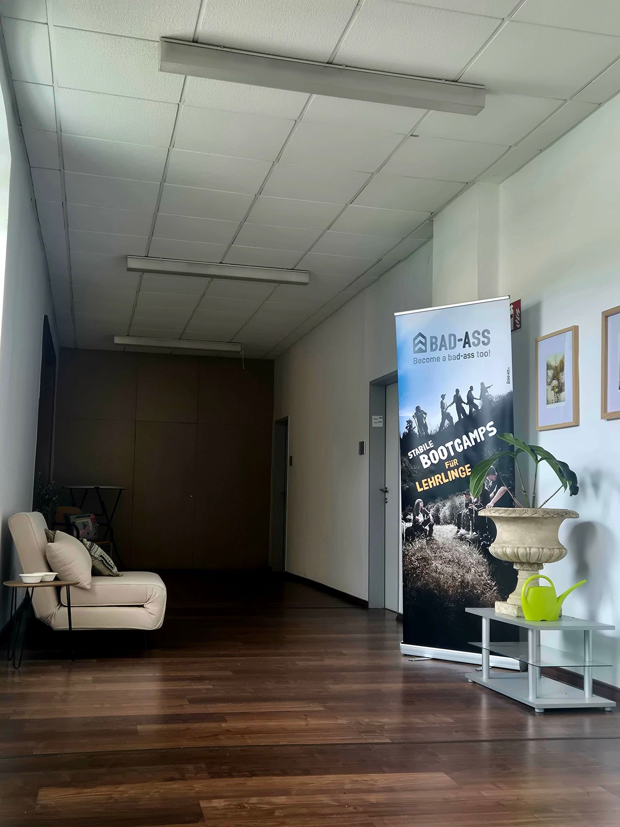 Ein leerer Flur mit einer weißen Couch, einem Tisch und einem Banner, das 'Stabile Bootcamps für Auszubildende' bewirbt. Es gibt eine Topfpflanze und einen Gießkannen auf einem Regal.