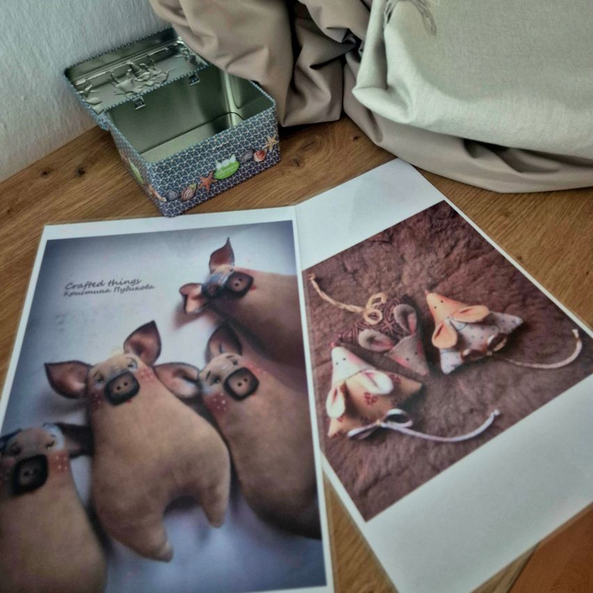 Auf einem Holztisch sind zwei gedruckte Fotos von Stofftieren zu sehen, eines mit drei Schweinen und das andere mit Mäusen, neben einer dekorativen Blechdose.