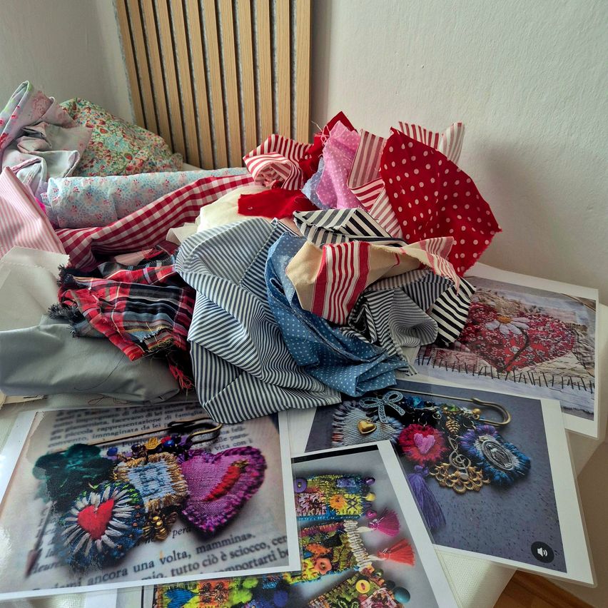 Ein Tisch mit verschiedenen farbigen Stoffresten, herzförmiger Stickerei und bedruckten Papieren. Die Stoffe sind in verschiedenen Farben und Mustern, und die herzförmige Stickerei ist auf weißem Hintergrund. Die bedruckten Papiere haben Bilder von Blumen, Herzen und anderen Designs.