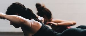 Eine Frau trainiert mit einer Hantel, liegt auf dem Boden und konzentriert sich auf ihr Workout. Eine weitere Frau ist im Hintergrund verschwommen, ebenfalls beim Training.