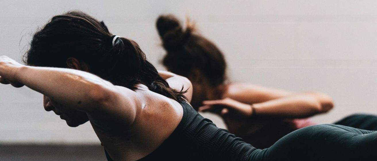 Eine Frau trainiert mit einer Hantel, liegt auf dem Boden und konzentriert sich auf ihr Workout. Eine weitere Frau ist im Hintergrund verschwommen, ebenfalls beim Training.