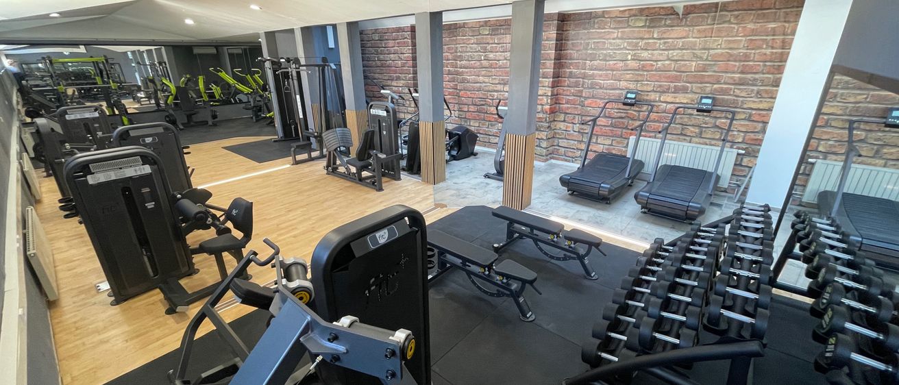 Ein Fitnessstudio mit verschiedenen Trainingsgeräten wie Laufbändern, Kraftmaschinen und Hanteln. Das Studio hat eine Ziegelwand auf einer Seite und einen Holzboden.