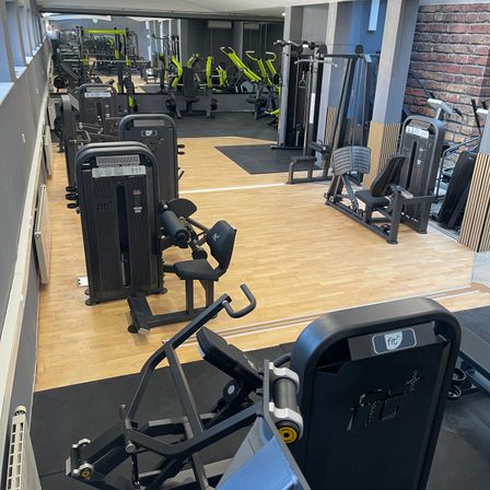 Der Fitnessraum ist mit verschiedenen Fitnessgeräten wie Kraftmaschinen, Laufbändern und Bankdrücken ausgestattet. Der Boden des Fitnessraums besteht aus Holz, und die Wände sind aus Ziegeln.