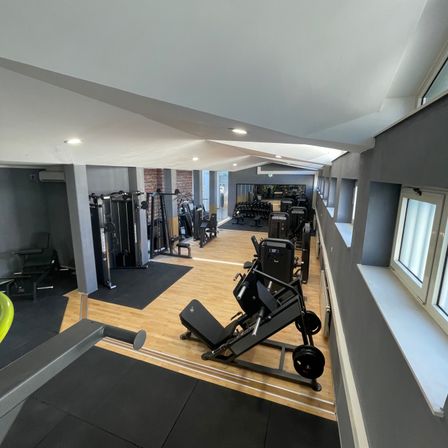 Innengym mit verschiedenen Trainingsgeräten, Holzböden und großen Fenstern.