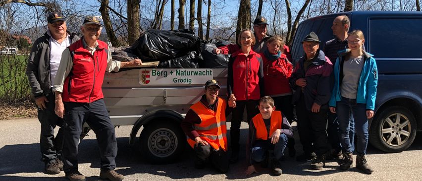 Eine Gruppe von Menschen in roten Westen posiert für ein Foto vor einem Anhänger mit einem Banner, das 'Berg + Naturwacht Grodig' besagt.