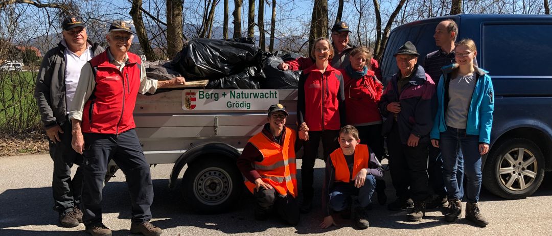 Eine Gruppe von Menschen in roten Westen posiert für ein Foto vor einem Anhänger mit einem Banner, das 'Berg + Naturwacht Grodig' besagt.