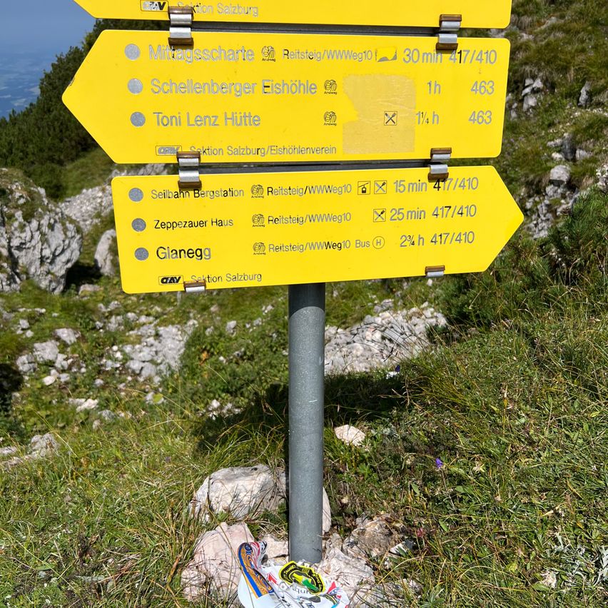 Ein gelbes Wanderschild zeigt Richtungen und Zeiten für verschiedene Orte an. Es weist einen 30-minütigen Weg zu Mittagscharte, 1 Stunde zu Schellenberger Eishöhle und 1,5 Stunden zu Toni Lenz Hütte aus. Ein weiteres Schild gibt 15 Minuten, 25 Minuten und 2,5 Stunden für Seilbahn Bergstation, Zeppezauer Haus und Glanegg an.