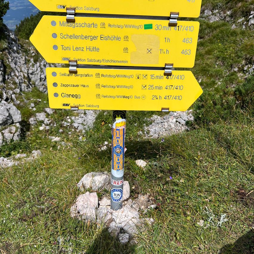Ein gelber Wegweiser mit Informationen zu Wanderwegen. Es zeigt Ziele wie Schellenberger Eishöhle und Toni Lenz Hütte. Zeitschätzungen sind für jedes Ziel angegeben.