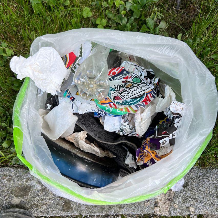 Ein durchsichtiger Plastikbeutel ist voller Müll, einschließlich Plastikflaschen, Papier und Lebensmittelverpackungen, auf dem Boden in der Nähe von Gras platziert.