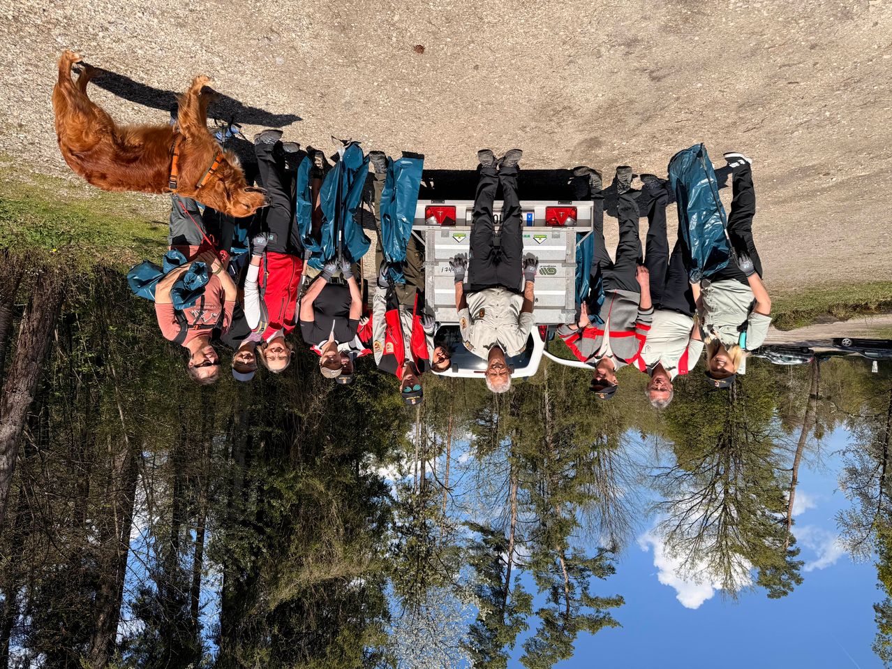 A group of people and a dog are standing in a line with their arms raised. They are next to a vehicle and surrounded by trees. The image has a reflection effect.