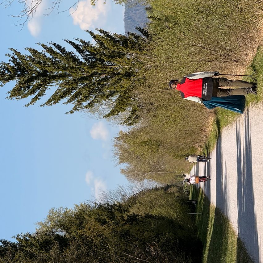 A few people are riding bicycles on a gravel road. One person in the foreground wears a red vest and holds a cane. Trees line the roadside.