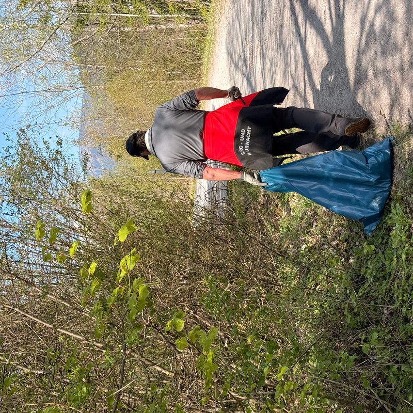 A man wearing a red and gray shirt is picking up trash from the roadside with a blue bag.