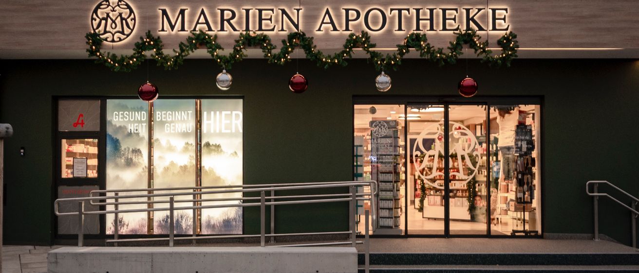 Die Fassade der Marien Apotheke zeigt ein Logo, festliche Dekorationen und eine Glastür mit Eingangsschild. Der Laden präsentiert Produkte im Inneren, mit einem Handlauf für Barrierefreiheit.