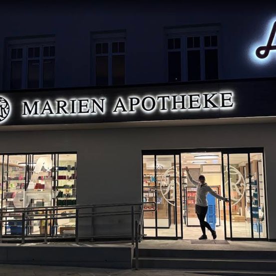 Eine Person steht nachts vor dem Eingang einer Apotheke. Der Laden heißt Marien Apotheke.