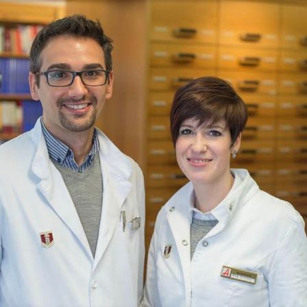 Ein Mann und eine Frau in weißen Laborkittel lächeln für die Kamera vor einem hölzernen Bücherregal.