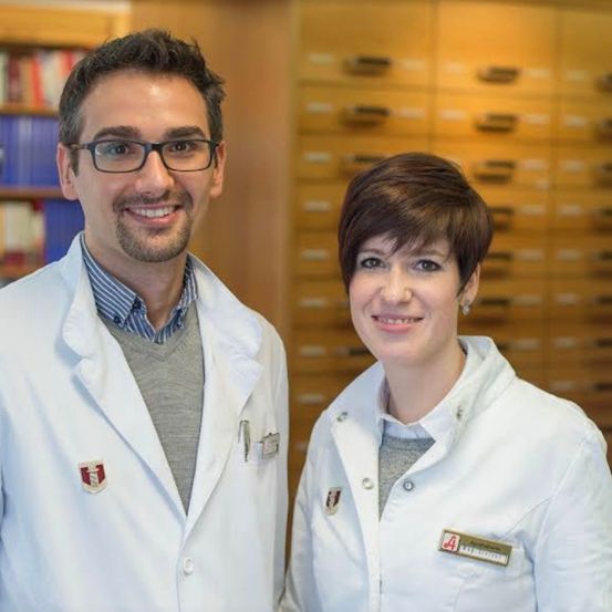 Ein Mann und eine Frau in weißen Laborkittel lächeln für die Kamera vor einem hölzernen Bücherregal.