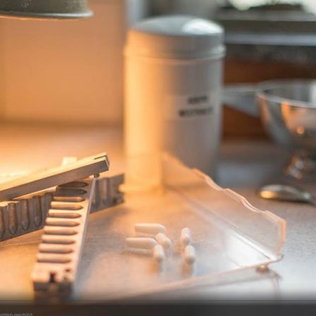 Ein Tisch mit einem Metallobjekt und mehreren weißen Pillen auf einem durchsichtigen Tablett. Ein weißer Krug und eine Schüssel sind im Hintergrund.