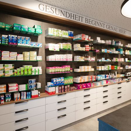Ein Innenbereich einer Apotheke mit einer Wand, die den Text Gesundheit beginnt genau hier! trägt. Die Regale sind mit Boxen von Medikamenten und anderen Produkten gefüllt.
