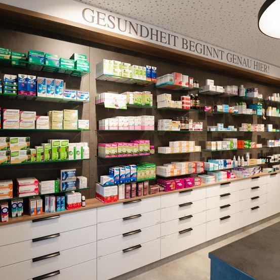Ein Innenbereich einer Apotheke mit einer Wand, die den Text Gesundheit beginnt genau hier! trägt. Die Regale sind mit Boxen von Medikamenten und anderen Produkten gefüllt.