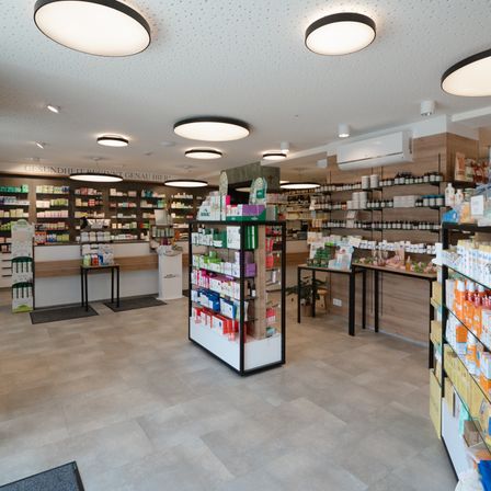 Das Innere einer Apotheke mit gefliestem Boden, modernen Deckenleuchten und ordentlich geordneten Regalen, die mit verschiedenen Produkten bestückt sind. Hinter den Regalen befindet sich eine Theke mit einem Topfpflanzen.