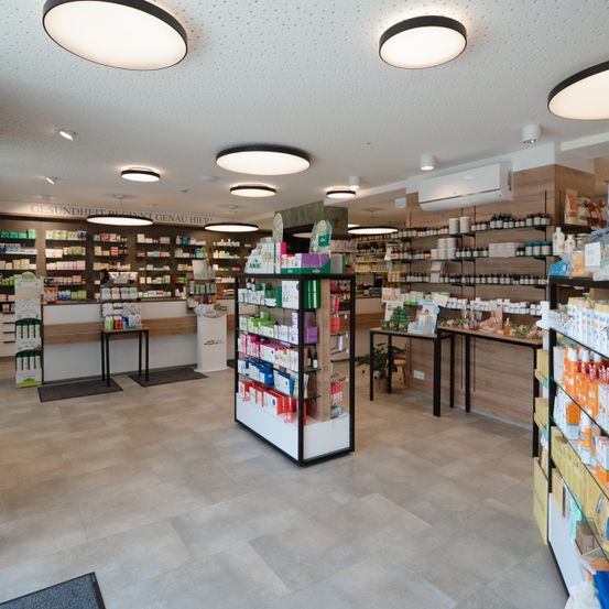 Das Innere einer Apotheke mit gefliestem Boden, modernen Deckenleuchten und ordentlich geordneten Regalen, die mit verschiedenen Produkten bestückt sind. Hinter den Regalen befindet sich eine Theke mit einem Topfpflanzen.