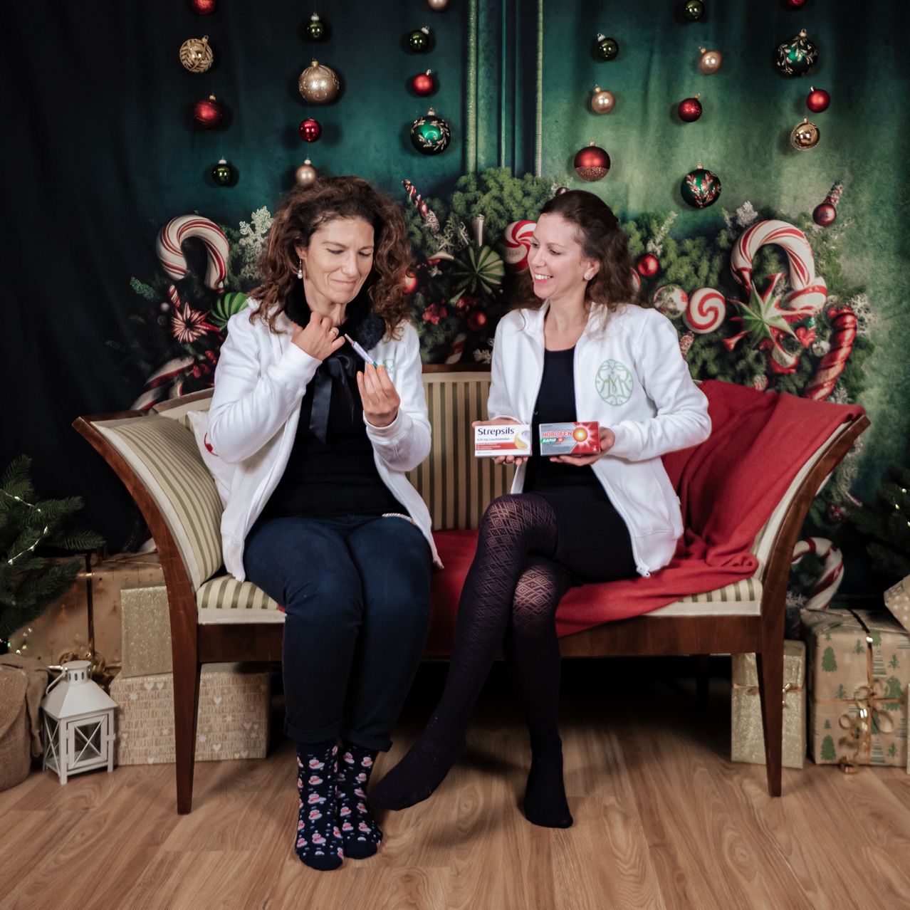 Zwei Frauen in Laborkittel sitzen auf einer Couch in einer festlichen Umgebung, lächelnd und halten eine Schachtel Strepsils. Sie sind von Weihnachtsdekorationen und Geschenken auf dem Holzboden umgeben.