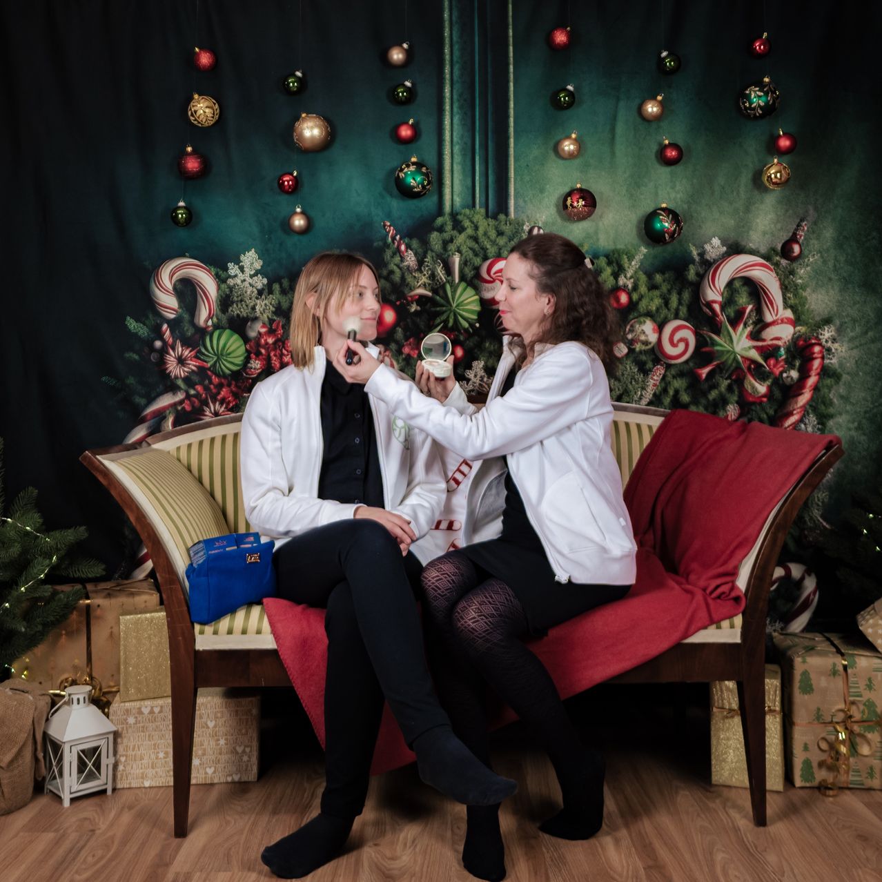 Zwei Frauen in weißen Laborkittel sitzen auf einer Couch, eine schminkt die andere. Sie sind von Weihnachtsdekorationen umgeben, einschließlich Ornamenten und Zuckerstangen. Geschenke sind im Raum verstreut.
