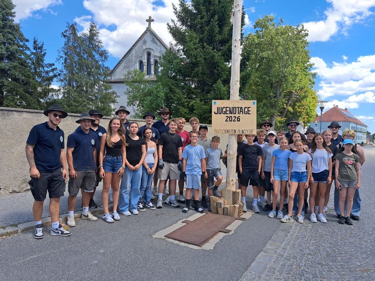 Eine Gruppe von Menschen posiert für ein Foto vor einem Schild, das Jugendtage 2026 sagt. Sie sind am Straßenrand.