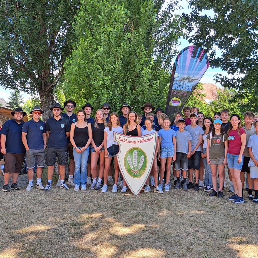 Eine Gruppe von Menschen, möglicherweise ein Team, steht vor Bäumen mit einem Banner im Hintergrund. Sie lächeln und posieren für ein Gruppenfoto.