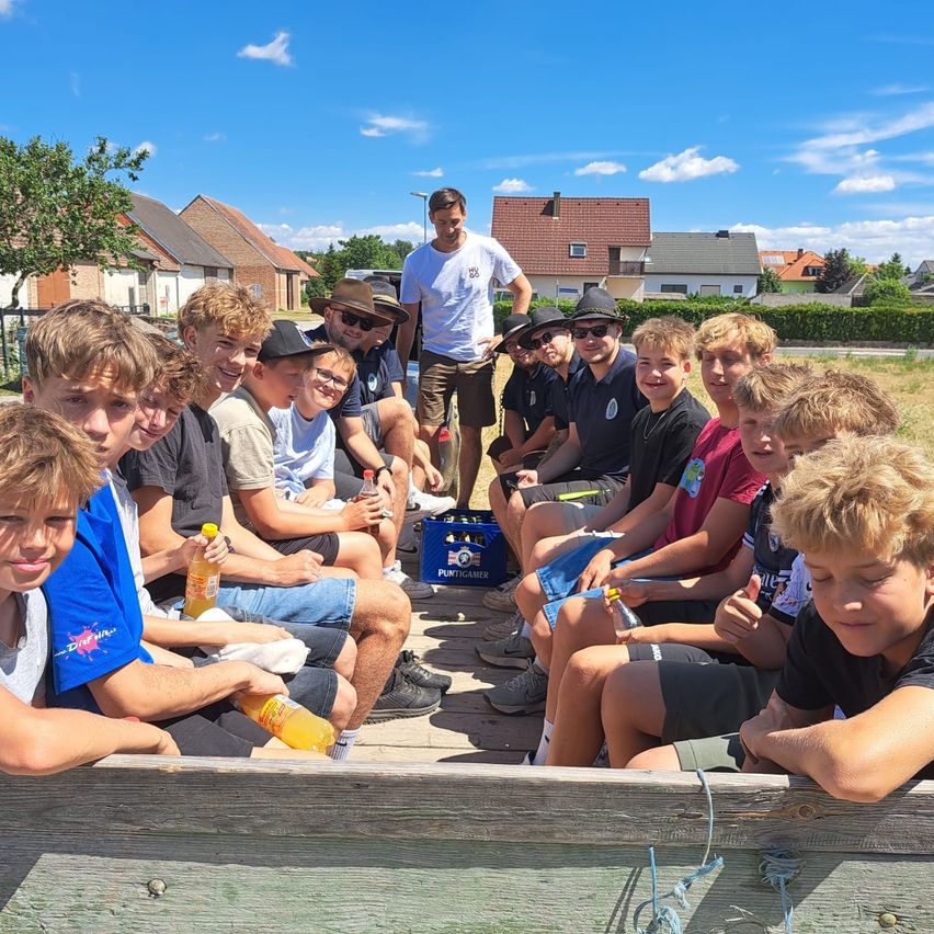 Eine Gruppe junger Männer sitzt zusammen auf einem Anhänger, hinter ihnen steht ein Mann. Sie befinden sich in einer sonnigen Umgebung im Freien mit Häusern und Bäumen im Hintergrund.