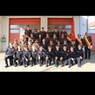 Eine Gruppenaufnahme von Feuerwehrleuten, sowohl Erwachsenen als auch Kindern, wird vor einem roten Feuerwehrhaus gemacht. Sie sind alle in Uniformen gekleidet.