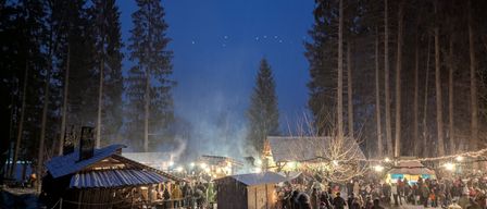Ein schneeiger Nachtmarkt im Wald, wo Menschen Essen und Getränke genießen, umgeben von beleuchteten Hütten und Bäumen unter einem blauen Himmel.