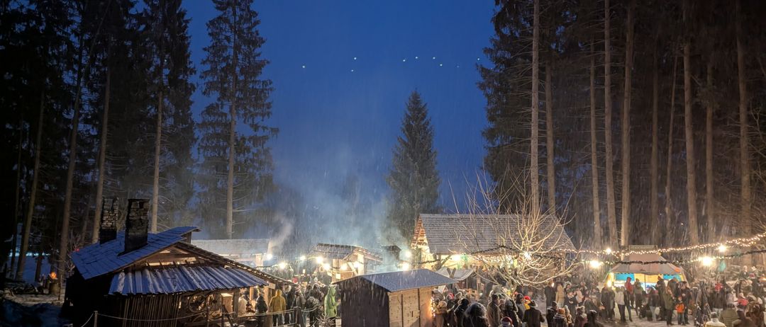 Ein schneeiger Nachtmarkt im Wald, wo Menschen Essen und Getränke genießen, umgeben von beleuchteten Hütten und Bäumen unter einem blauen Himmel.