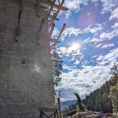 Eine Steinmauer mit Holzgerüst unter einem blauen Himmel mit Wolken, beleuchtet von Sonnenlicht. Bäume und Berge im Hintergrund.