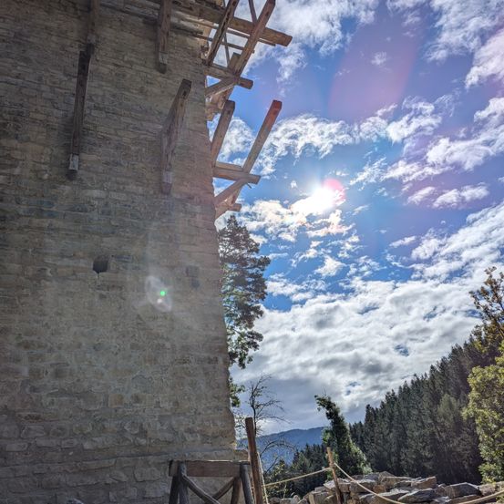 Eine Steinmauer mit Holzgerüst unter einem blauen Himmel mit Wolken, beleuchtet von Sonnenlicht. Bäume und Berge im Hintergrund.