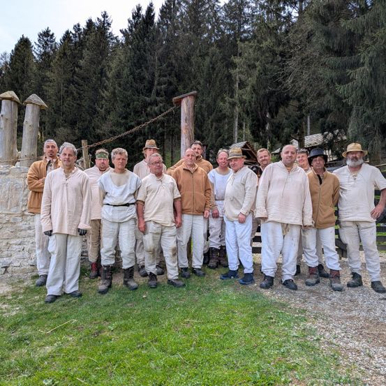 Eine Gruppe von Männern in weißen und beigefarbenen Outfits steht in einer ländlichen Umgebung. Sie tragen Hüte und Handschuhe. Hinter ihnen ist eine Steinmauer und eine Holzstruktur mit einer Kette. Der Hintergrund ist mit hohen Bäumen gefüllt.