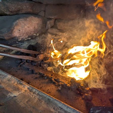Ein Paar Zangen hält brennende Kohlen in einem Steinkamin fest, mit hellen Flammen und aufsteigendem Rauch.