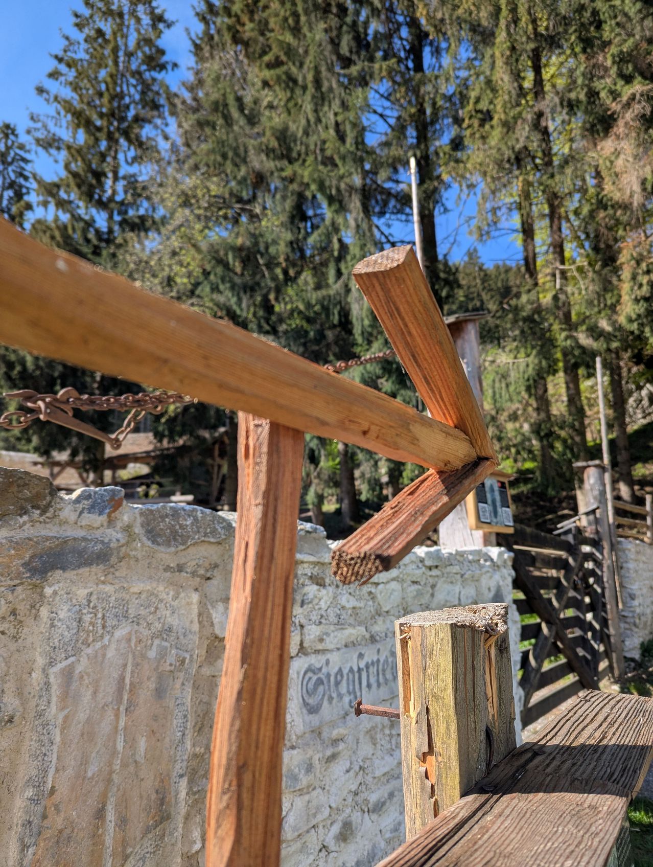Ein gebrochener Holzpfosten lehnt an einer Steinmauer mit dem Schild Siegfried. Im Hintergrund sind Bäume und ein Tor zu sehen.
