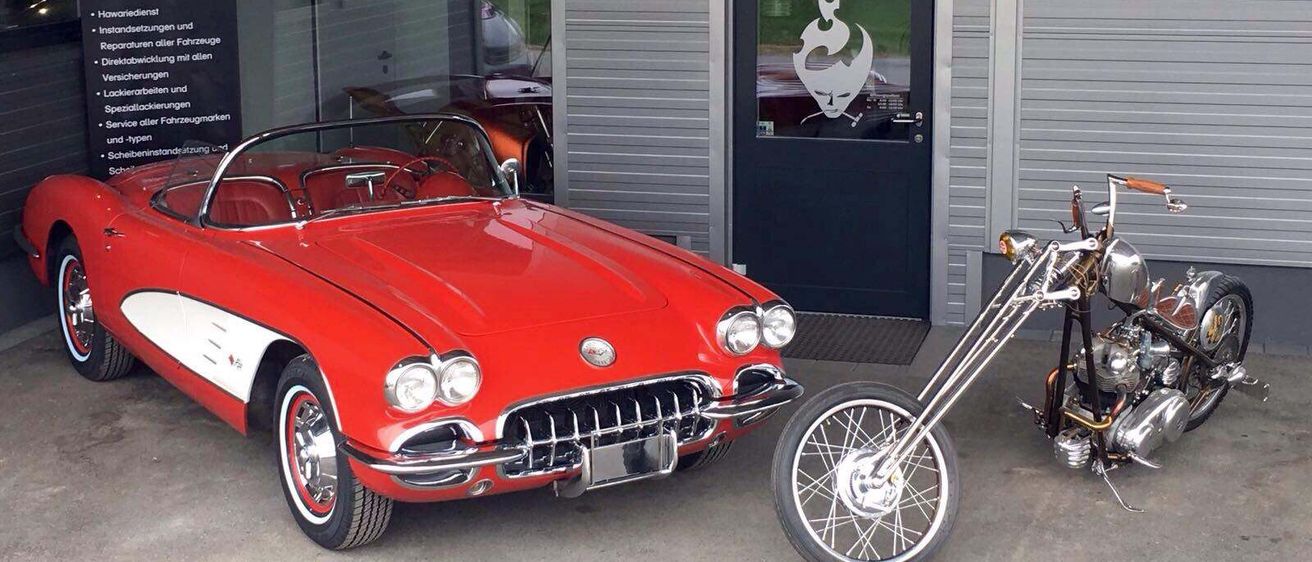 Ein roter Vintage Chevrolet Corvette Cabriolet ist neben einem Motorrad vor einem grauen Gebäude mit einer Glastür geparkt.